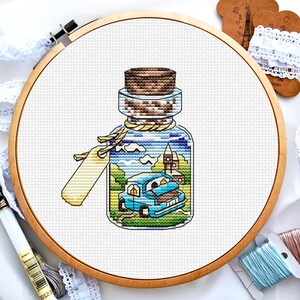 May include: A cross-stitch embroidery of a glass bottle with a cork stopper. The bottle contains a miniature scene of a blue car driving through a town with a church and houses. The bottle is on a white background.