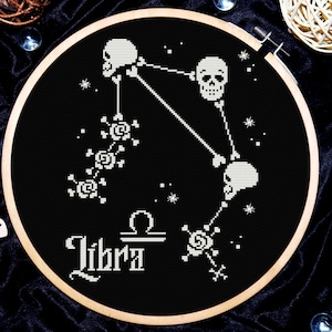 Puede incluir: Aro de bordado de punto de cruz en blanco y negro con un diseño de calavera y rosa que representa el signo del zodíaco Libra. El texto "Libra" está bordado debajo del diseño.