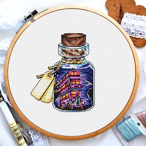Könnte beinhalten: Kreuzstich-Kunst einer Glasflasche mit Korken, die eine farbenfrohe Illustration eines Doppeldeckerbusses enthält. Die Flasche ist mit Bindfaden und einem leeren Etikett gebunden. Das Kunstwerk befindet sich in einem hölzernen Stickrahmen.