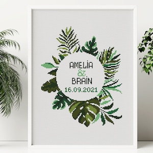 May include: Cross stitch pattern featuring a green tropical leaf wreath with a circle in the center. The circle contains the text "Amelia & Brain 16.09.2021".
