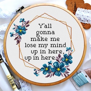 May include: A white embroidery hoop with a wooden frame, featuring a cross-stitch design of a gold geometric frame with blue and purple flowers and the text "Y'all gonna make me lose my mind up in here, up in here".