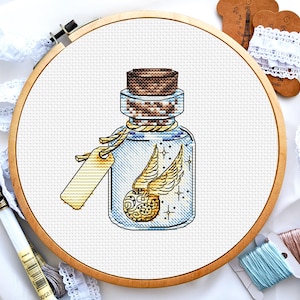 May include: A cross-stitch pattern of a glass bottle with a cork stopper. The bottle contains a golden winged object and sparkles. The bottle has a tag attached to it.