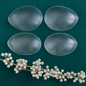 May include: Four clear silicone bra inserts on a green background. A branch with white pearl-like berries is in the bottom center of the image.
