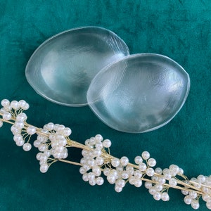May include: Two clear silicone bra inserts on a green background. The inserts are shaped like half circles and have a textured surface.
