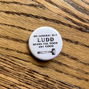 Peut inclure: Bouton blanc avec du texte noir qui dit "No general but Ludd means the poor any good" avec un dessin de marteau.