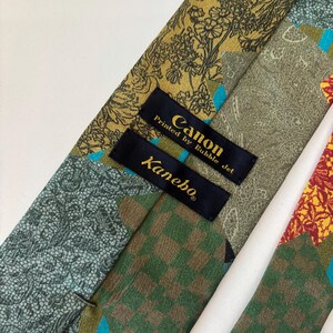 May include: Close-up of a patterned necktie with various designs and colors. The tie features floral, paisley, and checkered patterns in shades of green, gold, and red. The label reads "Canon Printed by Bubble Jet" and "Kanebo".