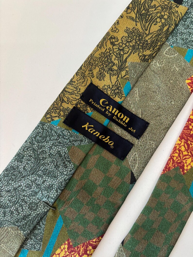 May include: Close-up of a patterned necktie with various designs and colors. The tie features floral, paisley, and checkered patterns in shades of green, gold, and red. The label reads "Canon Printed by Bubble Jet" and "Kanebo".