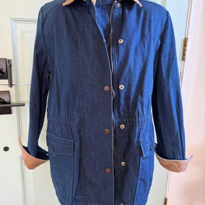 May include: A blue denim jacket with a brown corduroy collar and cuffs. The jacket has a zipper closure, button details, and two front pockets. It features a drawstring waist and is displayed on a mannequin.