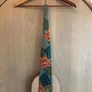 May include: A patterned necktie with a mix of green, red, and black geometric designs. The tie is hanging on a wooden hanger against a light wood background. The tie's design includes squares and floral patterns.