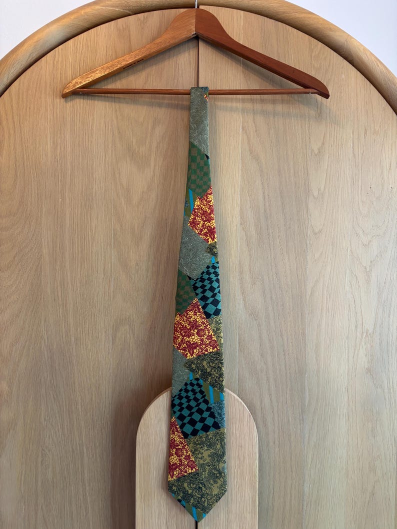 May include: A patterned necktie with a mix of green, red, and black geometric designs. The tie is hanging on a wooden hanger against a light wood background. The tie's design includes squares and floral patterns.