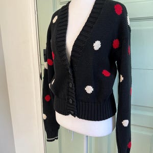 May include: Black cardigan sweater with a V-neck and button closure. The sweater features a pattern of red and white polka dots. The cuffs and hem have a white stripe detail. The sweater is displayed on a mannequin.