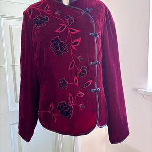 May include: A deep red velvet jacket featuring a mandarin collar and black trim. The front is decorated with black floral embroidery and three black toggle closures. The jacket has long sleeves and a structured fit, displayed on a mannequin.