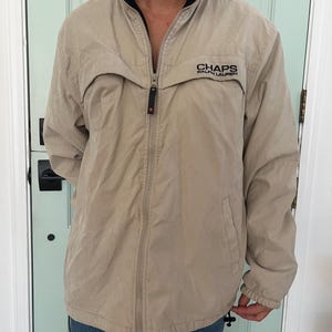 May include: Tan Chaps Ralph Lauren jacket with a black collar and a full zipper. The jacket has the brand name printed on the chest. The jacket is worn over blue jeans.