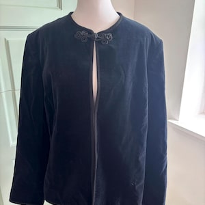 May include: A black velvet jacket featuring a round neckline and a single decorative frog closure at the top. The jacket has long sleeves, a straight hemline, and black piping trim along the front opening and edges, displayed on a mannequin.