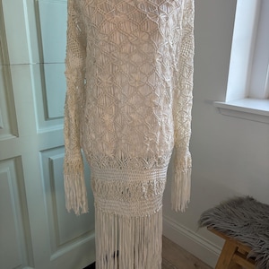 May include: An off-white long-sleeved dress featuring an intricate macrame knot pattern throughout the bodice and sleeves. The garment includes a wide, textured woven band at the waist and a long, straight fringe trim along the bottom hem.