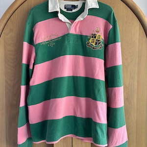 May include: A long-sleeve rugby shirt featuring wide horizontal stripes in green and pink. The shirt has a white collar, a button placket, and embroidered crests on the chest. Text on the chest reads Polo by Ralph Lauren and RL Team Championship.