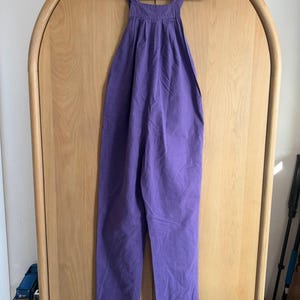 May include: A solid purple jumpsuit hangs on a white hanger against a light wood-grain cabinet. The jumpsuit features a square neckline, wide straps, and wide-leg pants. The fabric appears to be a lightweight material.