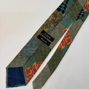 May include: A patterned necktie with a patchwork design. The tie features various patterns including floral, paisley, and checkered designs in shades of green, brown, red, and blue. The tie has a navy blue lining and labels that read "Canon" and "Kanebo".