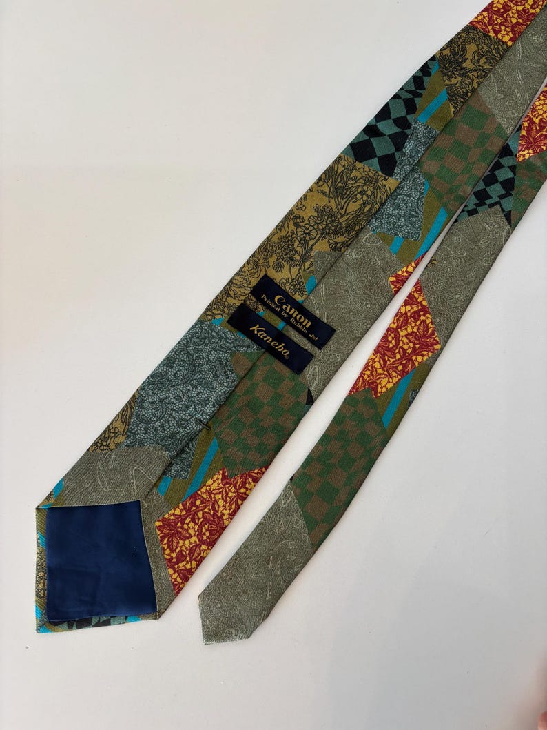 May include: A patterned necktie with a patchwork design. The tie features various patterns including floral, paisley, and checkered designs in shades of green, brown, red, and blue. The tie has a navy blue lining and labels that read "Canon" and "Kanebo".