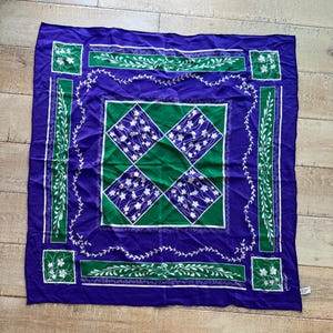 May include: A square, purple scarf with a green and white floral design. The scarf features a central green diamond pattern with white floral accents, framed by a purple border with green and white decorative elements. The scarf is made of a soft, flowing fabric.