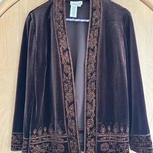 May include: A dark brown velvet jacket featuring an open front design with long sleeves. The garment is adorned with intricate copper-colored floral embroidery along the front placket, the bottom hem, and the cuffs. A white brand label is visible.