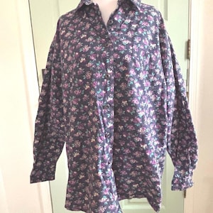 May include: A long-sleeved button-down shirt with a floral pattern. The shirt features a dark navy blue background with a repeating pattern of small purple, pink, and white flowers. The shirt is displayed on a mannequin.
