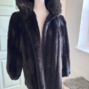 May include: A dark brown faux fur coat with a hood, displayed on a mannequin. The coat has a long, straight cut and button closures. The fur appears soft and plush, creating a luxurious look. The coat is a classic outerwear piece.