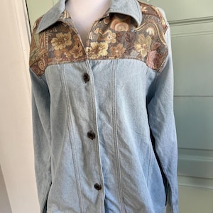 May include: Light blue denim jacket with a floral tapestry yoke and collar. The jacket has a button-up front and long sleeves. The floral pattern includes shades of brown, beige, and pink.