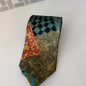 May include: A patchwork tie with various patterns and colors. The tie features sections of red floral, black and teal checkerboard, and olive green floral designs. The tie's tip has teal and blue stripes.