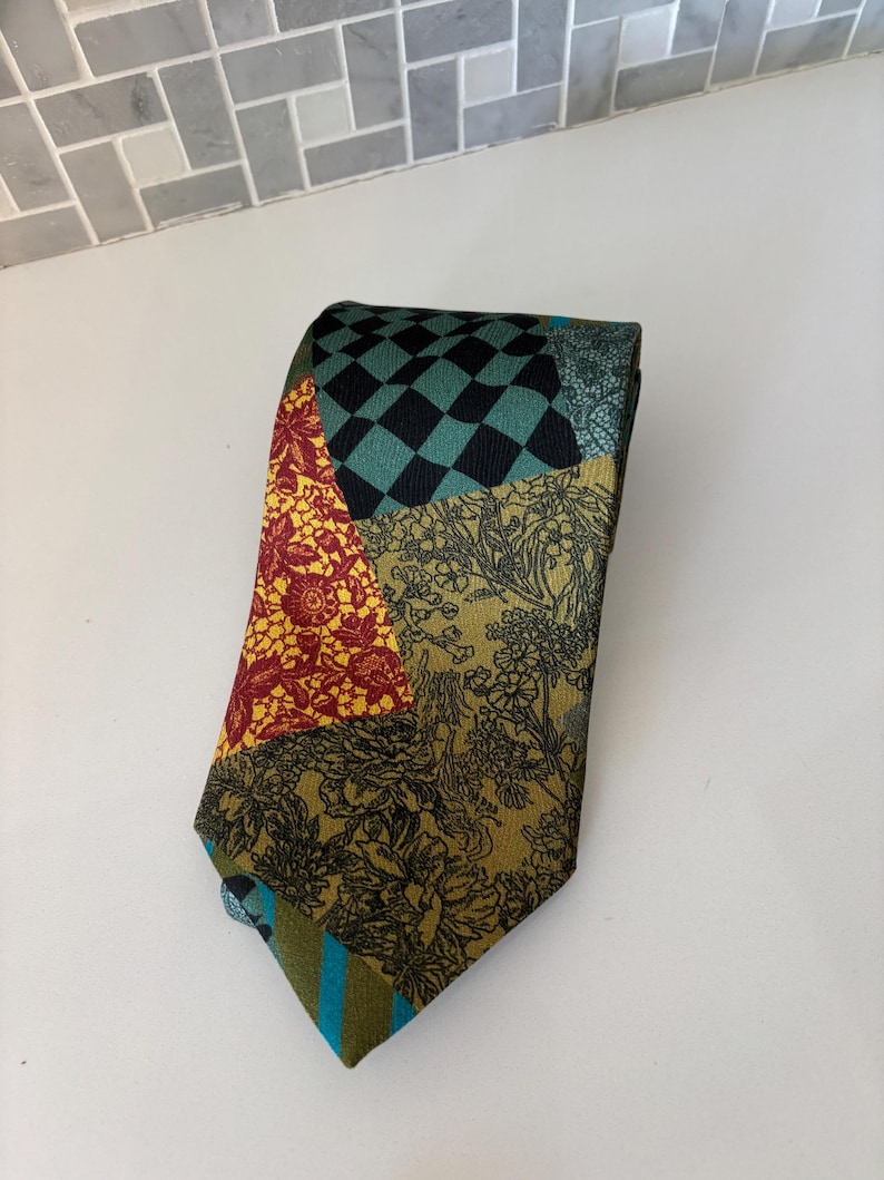 May include: A patchwork tie with various patterns and colors. The tie features sections of red floral, black and teal checkerboard, and olive green floral designs. The tie's tip has teal and blue stripes.
