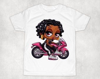 May include: A white t-shirt featuring a cartoon illustration of a Black girl with dreadlocks riding a pink motorbike. She is wearing a black and white outfit and white trainers.
