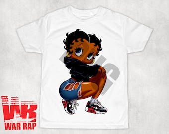 Op de afbeelding: Wit T-shirt met een cartoon afbeelding van een zwart vrouwelijk personage dat een zwarte hoodie, blauwe jeans en witte sneakers draagt. Het personage zit met gekruiste benen en heeft een speelse uitdrukking op haar gezicht.