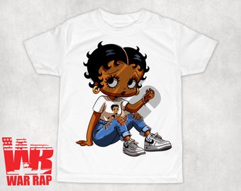 Op de afbeelding: Wit T-shirt met een cartoon afbeelding van een zwarte vrouw met bruin haar, die een blauwe jeans en witte sneakers draagt. De vrouw zit met gekruiste benen en heeft een klein beeld van zichzelf op haar shirt.