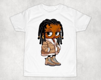 May include: White t-shirt featuring a cartoon illustration of a Black woman with long braids wearing a brown puffer jacket and brown and yellow trainers.