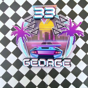 Puede incluir: Un adorno para pastel 3D azul y morado con un tema retro de los años 80. Presenta un coche deportivo azul, palmeras, una ciudad y el número 33. El nombre "GEORGE" está escrito en una fuente pixelada.