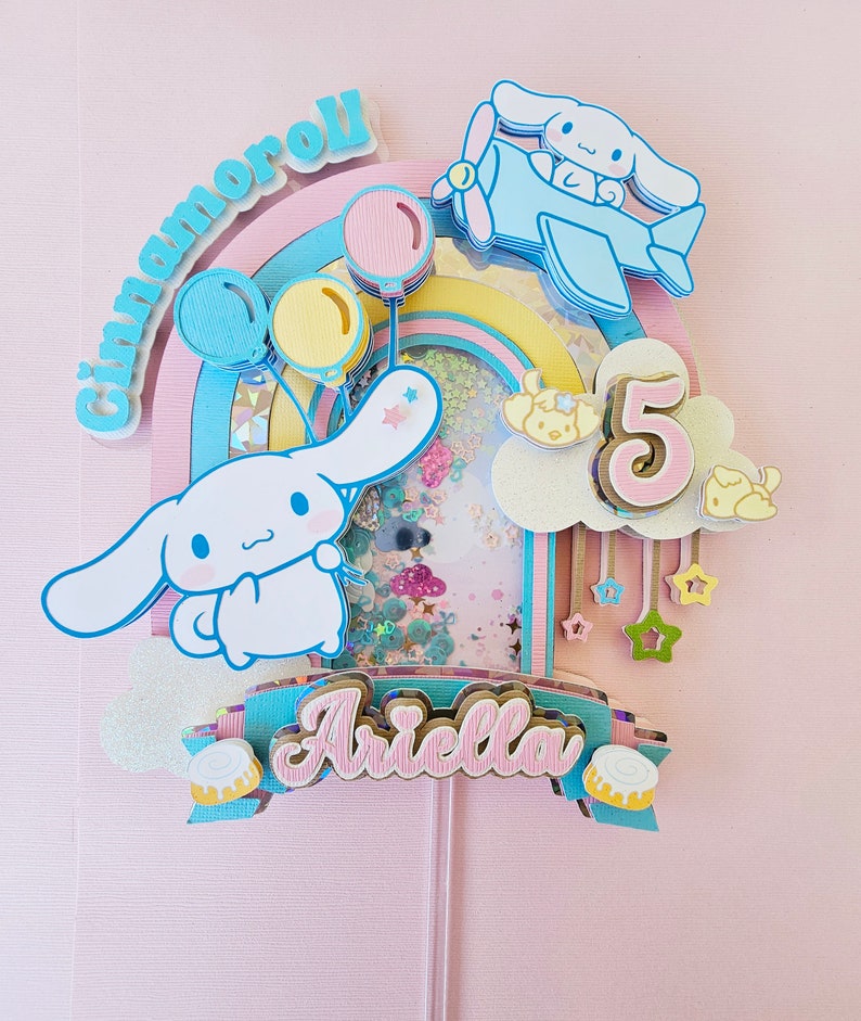 Pode incluir: Um topo de bolo rosa pastel, azul e amarelo com um arco-&iacute;ris, um personagem Cinnamoroll segurando bal&otilde;es, um avi&atilde;o com um personagem Cinnamoroll, uma nuvem com o n&uacute;mero 5 e uma faixa com o nome "Ariella".