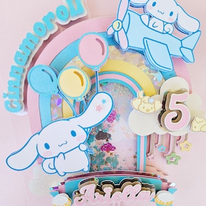 Pode incluir: Um topo de bolo rosa e azul com um arco-&iacute;ris, nuvens, estrelas e um personagem do desenho animado Cinnamoroll. O topo tamb&eacute;m inclui o nome "Ariella" e o n&uacute;mero "5".