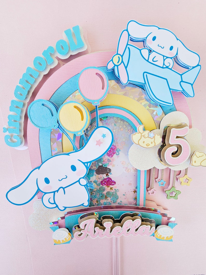 Pode incluir: Um topo de bolo rosa e azul com um arco-&iacute;ris, nuvens, estrelas e um personagem do desenho animado Cinnamoroll. O topo tamb&eacute;m inclui o nome "Ariella" e o n&uacute;mero "5".