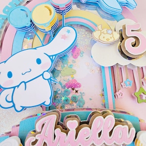 Pode incluir: Um topo de bolo arco-&iacute;ris rosa e azul com um personagem de cachorro branco, bal&otilde;es e o nome "Ariella" em letras rosa. O topo de bolo tamb&eacute;m inclui um n&uacute;mero "5" e um pequeno avi&atilde;o com um personagem de cachorro.