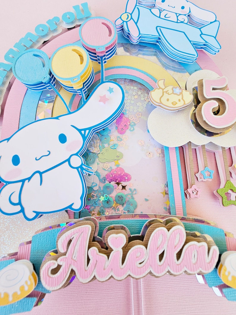 Pode incluir: Um topo de bolo arco-&iacute;ris rosa e azul com um personagem de cachorro branco, bal&otilde;es e o nome "Ariella" em letras rosa. O topo de bolo tamb&eacute;m inclui um n&uacute;mero "5" e um pequeno avi&atilde;o com um personagem de cachorro.