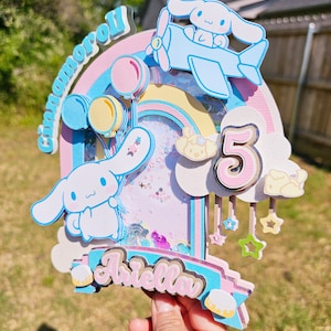 Pode incluir: Um topo de bolo em camadas rosa, azul e amarelo com um arco-&iacute;ris, nuvens, estrelas e um personagem Cinnamoroll voando em um avi&atilde;o azul. O topo de bolo tamb&eacute;m inclui o n&uacute;mero 5 e o nome "Ariella".