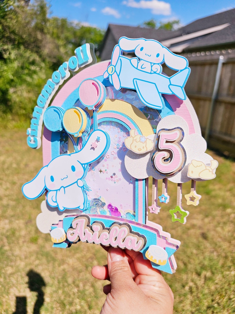 Pode incluir: Um topo de bolo em camadas rosa, azul e amarelo com um arco-&iacute;ris, nuvens, estrelas e um personagem Cinnamoroll voando em um avi&atilde;o azul. O topo de bolo tamb&eacute;m inclui o n&uacute;mero 5 e o nome "Ariella".