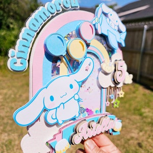 Pode incluir: Um topo de bolo rosa pastel, azul e branco com um arco-&iacute;ris, um personagem Cinnamoroll e o nome "Arella" em letras rosa. O topo tamb&eacute;m inclui um avi&atilde;o azul, bal&otilde;es e estrelas.