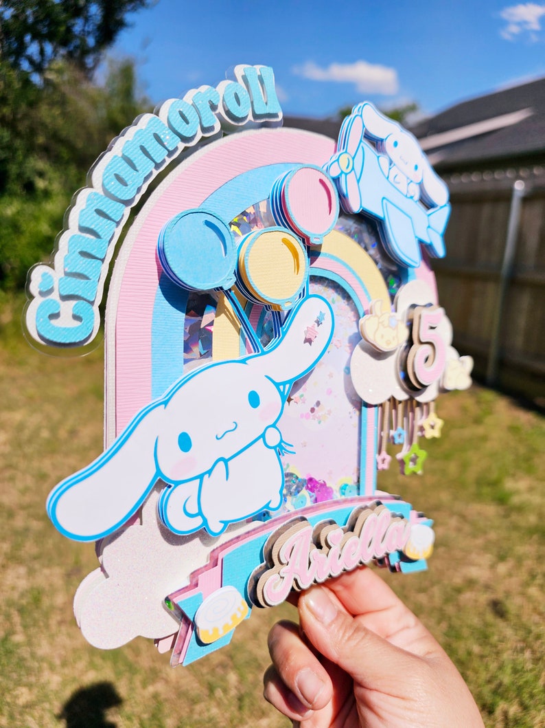 Pode incluir: Um topo de bolo rosa pastel, azul e branco com um arco-&iacute;ris, um personagem Cinnamoroll e o nome "Arella" em letras rosa. O topo tamb&eacute;m inclui um avi&atilde;o azul, bal&otilde;es e estrelas.