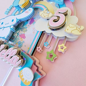 Pode incluir: Um topo de bolo rosa pastel com um arco-&iacute;ris, nuvens, estrelas e um personagem do desenho animado 'Hello Kitty'. O n&uacute;mero '5' est&aacute; no topo do bolo, junto com a palavra 'Bella'.