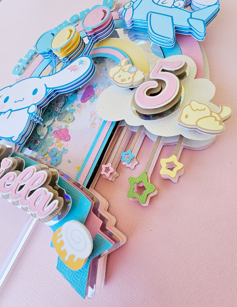 Pode incluir: Um topo de bolo rosa pastel com um arco-&iacute;ris, nuvens, estrelas e um personagem do desenho animado 'Hello Kitty'. O n&uacute;mero '5' est&aacute; no topo do bolo, junto com a palavra 'Bella'.
