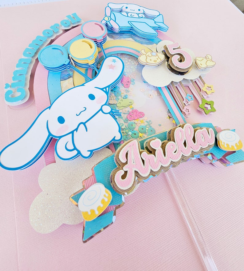 Pode incluir: Um topo de bolo com um personagem Cinnamoroll branco e rosa segurando bal&otilde;es, um arco-&iacute;ris, uma nuvem com o n&uacute;mero 5 e o nome Ariella em letras rosa.