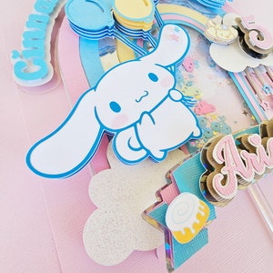 Pode incluir: Um topo de bolo rosa e azul com um c&atilde;o de desenho animado branco segurando bal&otilde;es. O c&atilde;o est&aacute; segurando um bal&atilde;o com a palavra "Cinnamoroll" nele. O topo de bolo tamb&eacute;m inclui um arco-&iacute;ris, uma nuvem e uma estrela.