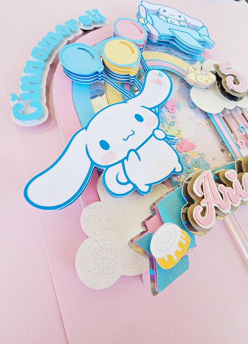 Pode incluir: Um topo de bolo rosa e azul com um c&atilde;o de desenho animado branco segurando bal&otilde;es. O c&atilde;o est&aacute; segurando um bal&atilde;o com a palavra "Cinnamoroll" nele. O topo de bolo tamb&eacute;m inclui um arco-&iacute;ris, uma nuvem e uma estrela.