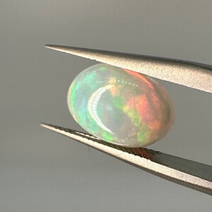 Puede incluir: Una piedra preciosa de ópalo blanco con destellos verdes, naranjas y rojos. El ópalo tiene forma ovalada y está sujeto con unas pinzas.